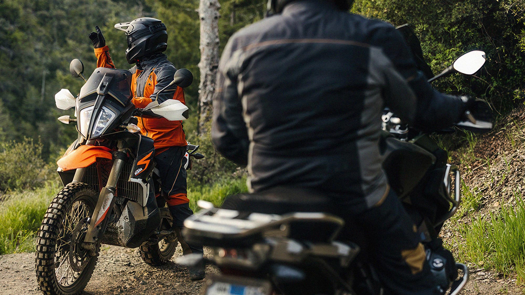Two adventure riders in textile jackets and adventure helmets on a forest gravel trail